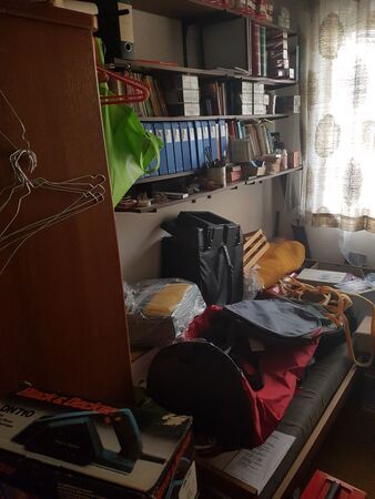 Kleines, vollgestelltes Arbeitszimmer mit Regalen, Aktenordnern und Kisten am Fenster.